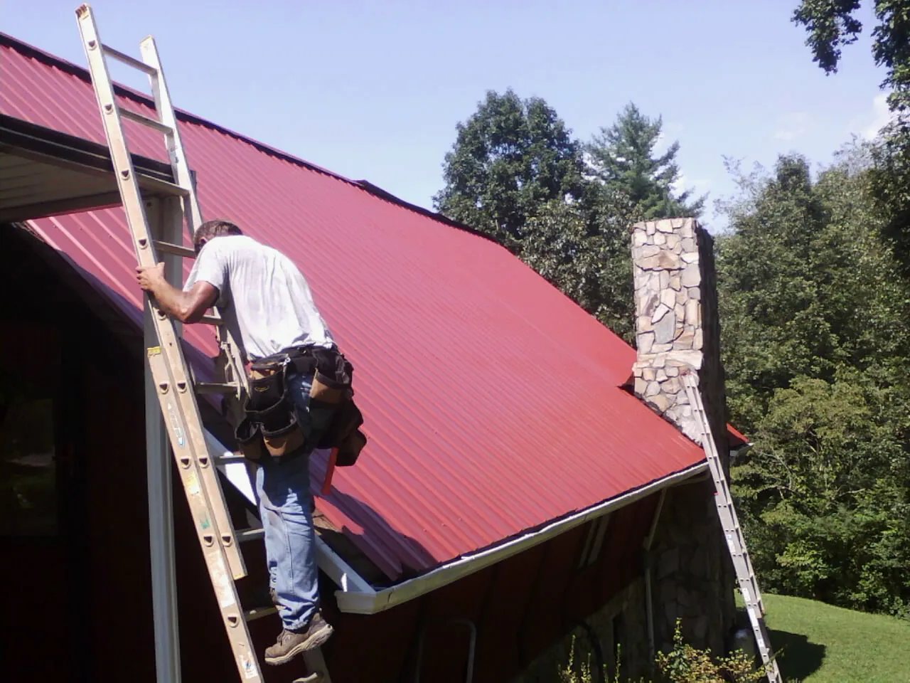 Roofer on a ladder beside a red metal roof and stone chimney — Soffit & Fascia Repair by Roofing Brothers in Widen